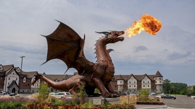 A giant cartoon-style dragon breathes fire over a landscaped hotel entrance with cars and buildings in the background.