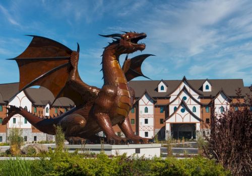 A large winged dragon statue roars in front of Tudor-style buildings under a blue sky.