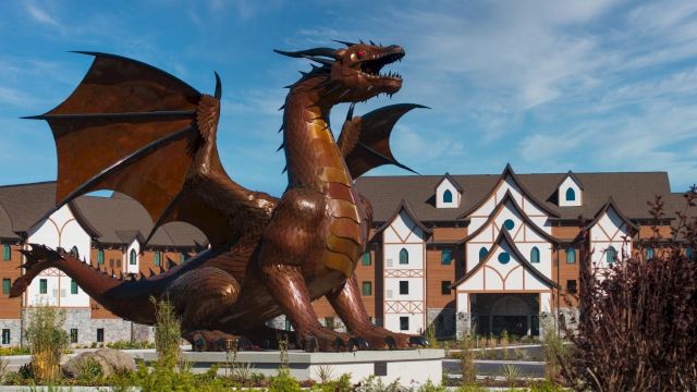 A large winged dragon statue roars in front of Tudor-style buildings under a blue sky.