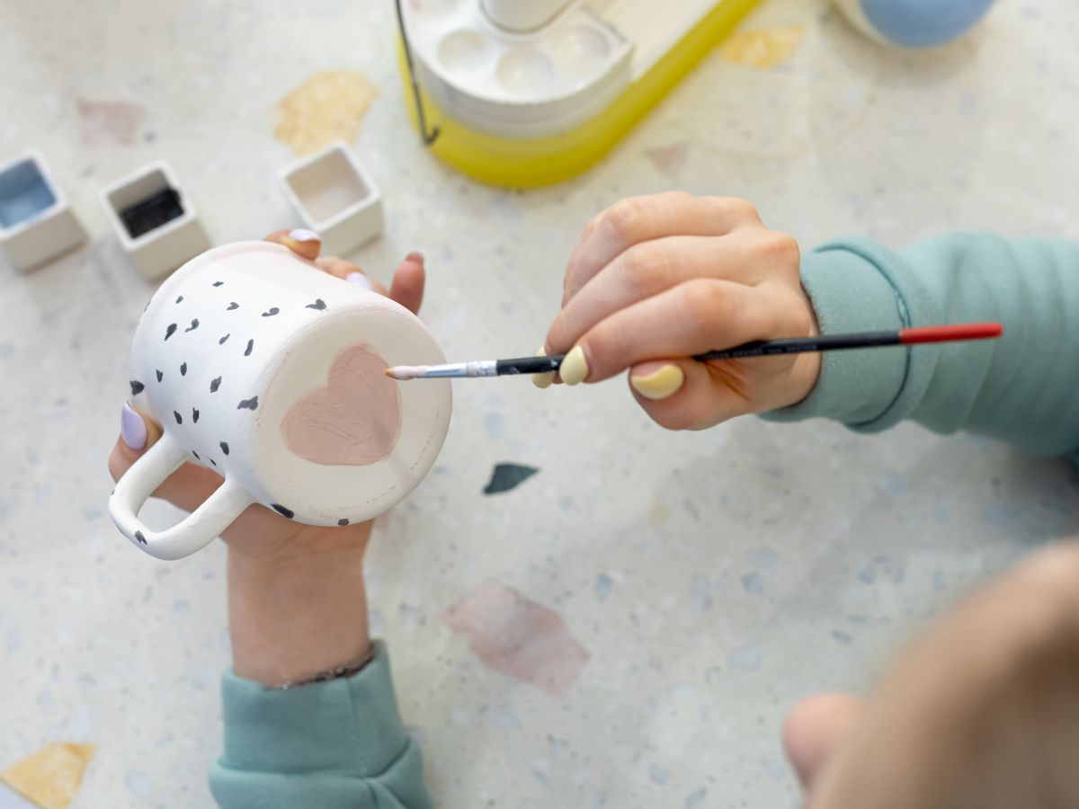 A person decorates a white ceramic mug with a paintbrush while craft supplies sit nearby.