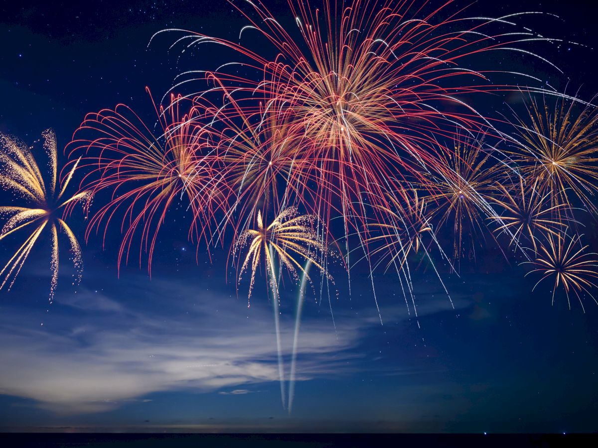 A nighttime sky filled with colorful fireworks&mdash;gold bursts, red streaks, and white sparks spreading over clouds.