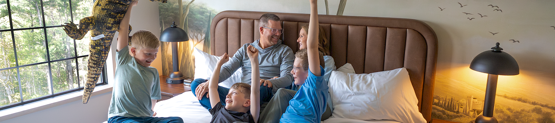 In a safari-themed bedroom, a dad and kids play and jump on a bed while a dinosaur-costumed child reaches up.
