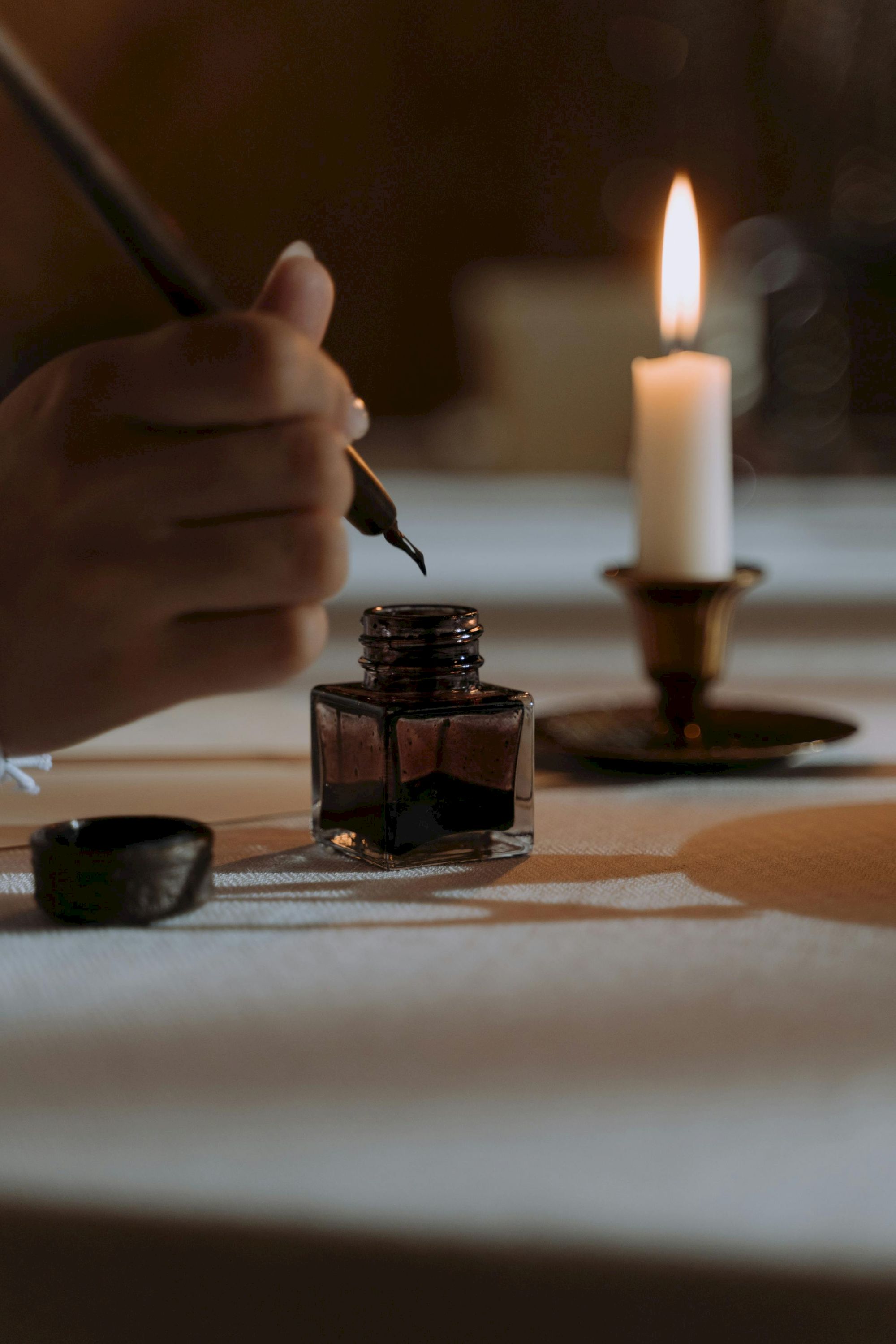 A hand dips a fountain pen into an ink bottle as a candle burns in the background.