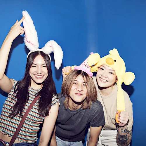 A group of friends pose with cute bunny and duck plush headbands on a blue backdrop, laughing together.