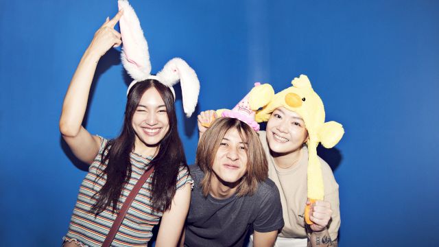 A group of friends pose with cute bunny and duck plush headbands on a blue backdrop, laughing together.