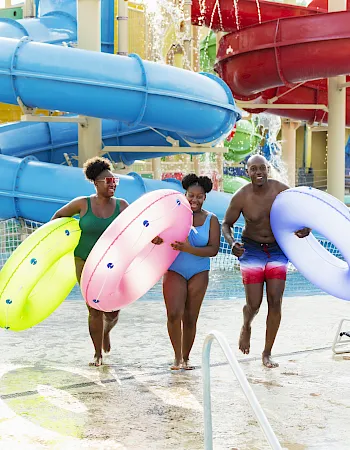 Smiling swimmers wade through a pool at a water park, holding colorful inner tubes as water sprays from giant slides.