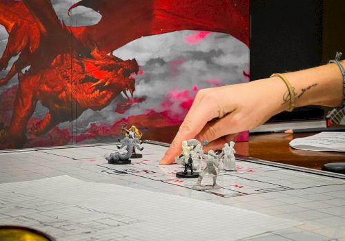 A tabletop battle scene with painted mini figures on a grid mat, a red dragon backdrop, and a hand positioning pieces.