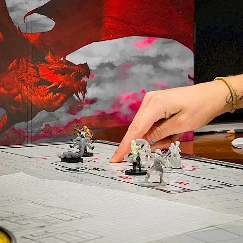 A tabletop battle scene with painted mini figures on a grid mat, a red dragon backdrop, and a hand positioning pieces.