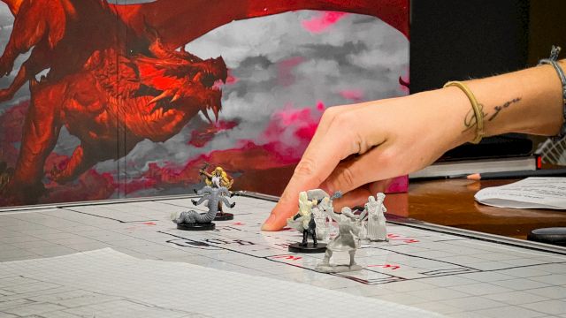 A tabletop battle scene with painted mini figures on a grid mat, a red dragon backdrop, and a hand positioning pieces.