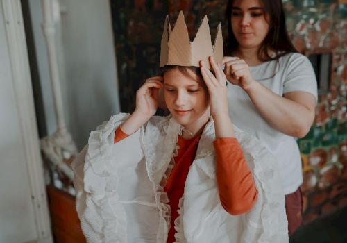 A child in a paper crown is smiling as an adult playfully helps adjust it, in a cozy home setting.
