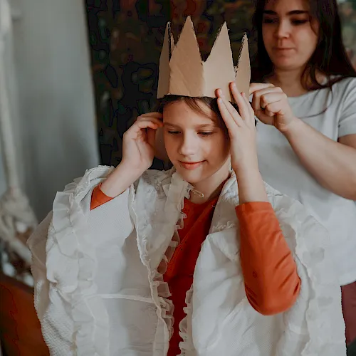 A child in a paper crown is smiling as an adult playfully helps adjust it, in a cozy home setting.