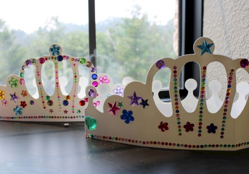 A pair of glittery gem-studded cardboard crowns with stars and colorful rhinestones sit by a window.