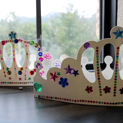 A pair of glittery gem-studded cardboard crowns with stars and colorful rhinestones sit by a window.