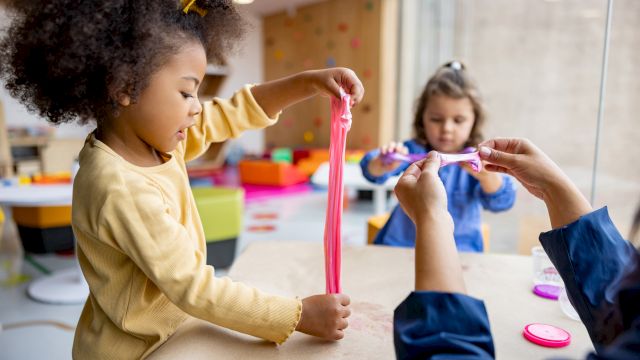 A child in a daycare plays with stretchy pink slime, holding it up while another child watches in the background.