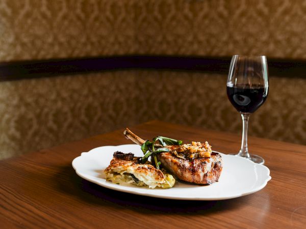 A plated meat entr&eacute;e with greens and a glass of red wine on a wooden table, ready for a cozy dinner.