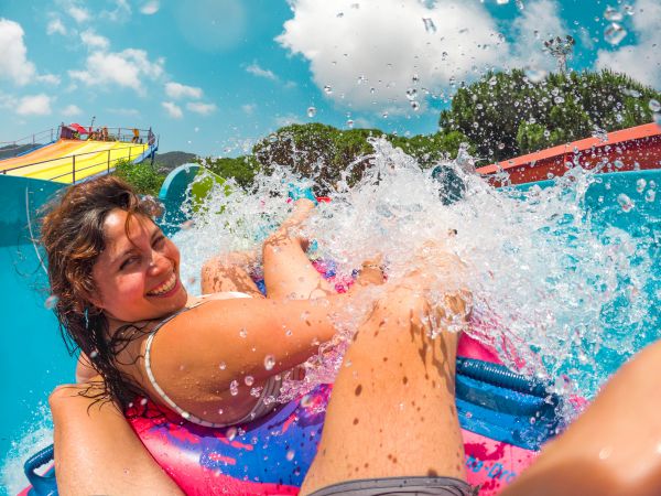 A couple slides into a sunny splash pool, water bursting around them at a water park as they grin in the spray.