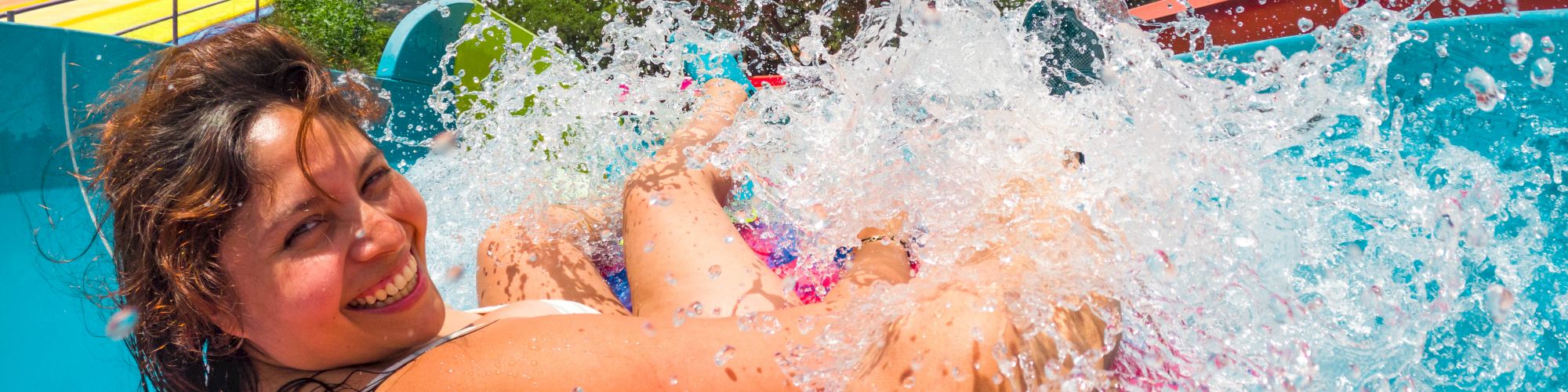 A water-park moment: a smiling woman gets soaked as a splash bursts from a water slide on a sunny day.