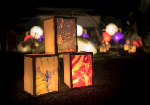 Colorful illuminated lantern boxes with painted scenes glow at night in a dark outdoor walkway.