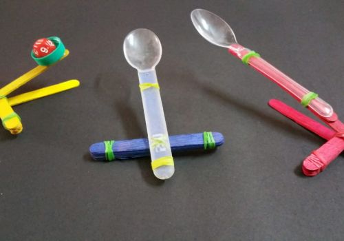 Three DIY maracas and shakers made from plastic spoons and bottle caps in bright yellow, blue, and red.