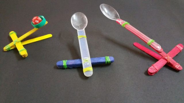 Three DIY maracas and shakers made from plastic spoons and bottle caps in bright yellow, blue, and red.