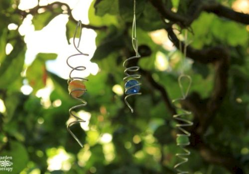 Two twisted metal hanging ornaments with small beads sway among green garden leaves in warm sunlight.