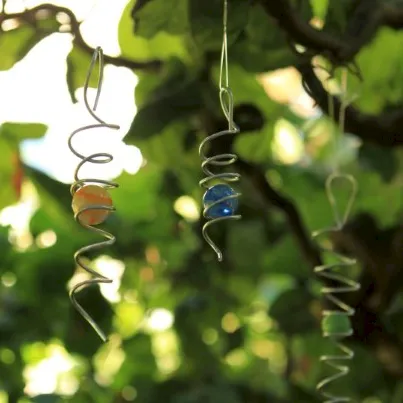 Two twisted metal hanging ornaments with small beads sway among green garden leaves in warm sunlight.