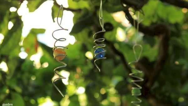 Two twisted metal hanging ornaments with small beads sway among green garden leaves in warm sunlight.
