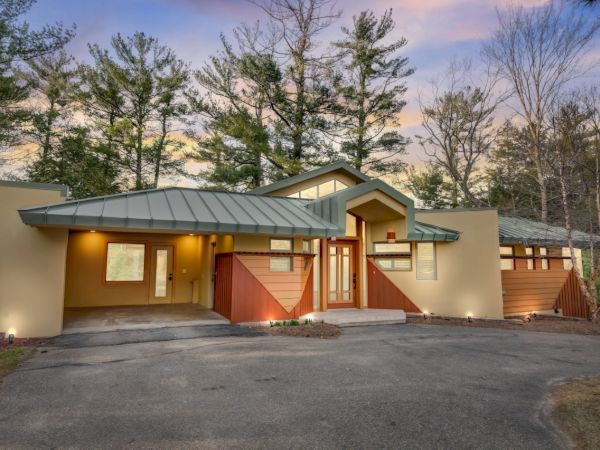 A modern home exterior with a driveway, red-brown doors, green metal roof, and tall trees against a sunset mountain backdrop.