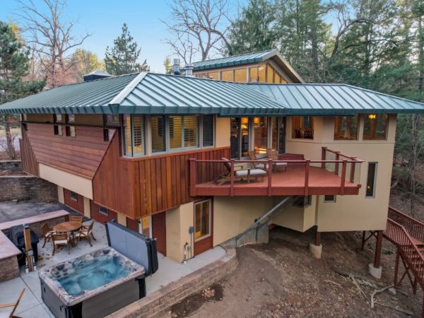 An outdoor view of a modern cabin with a covered deck, stairs, and a hot tub in a wooded area.
