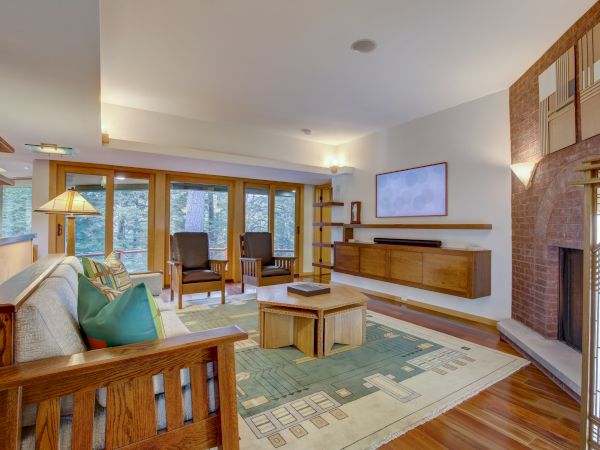 A cozy living room with a fireplace, wooden walls, large windows to trees, chairs, a sofa, and a patterned rug.