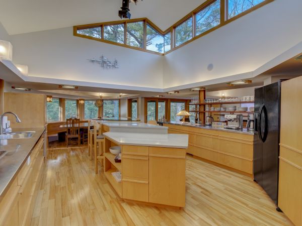 A bright modern kitchen with wood cabinets, large windows, stainless appliances, a built-in bar counter, and hanging glassware.