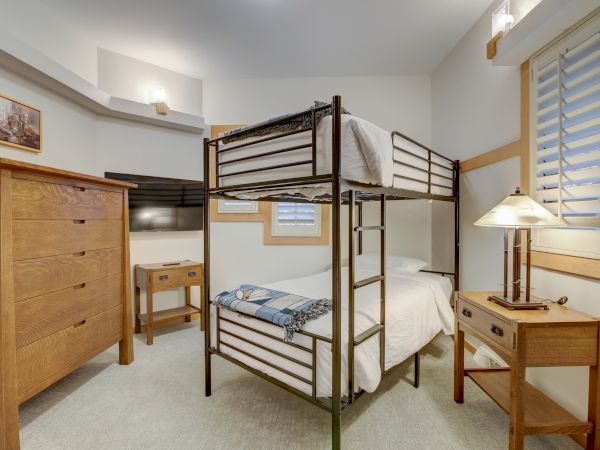 A tidy bedroom with a metal bunk bed, white bedding, bedside dresser, table lamp, and a window with blinds.