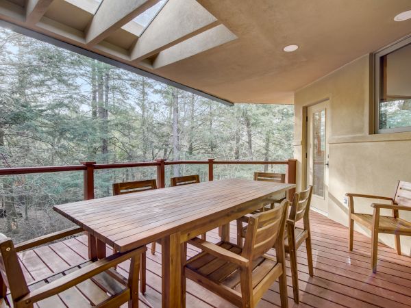 A covered wooden patio with a large table and chairs, glass doors, and forest trees visible through tall windows.