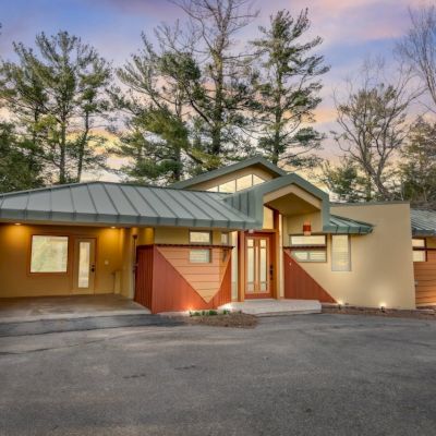 A modern home with a metal green roof, warm entry, and a wide driveway, surrounded by pine trees under a sunset sky.