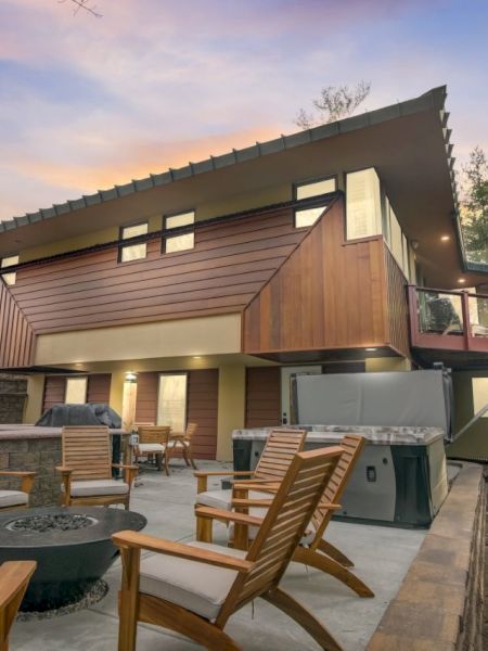 A modern cabin patio at dusk with deck chairs, a hot tub, and a green metal grill in front of a wood-sided house.