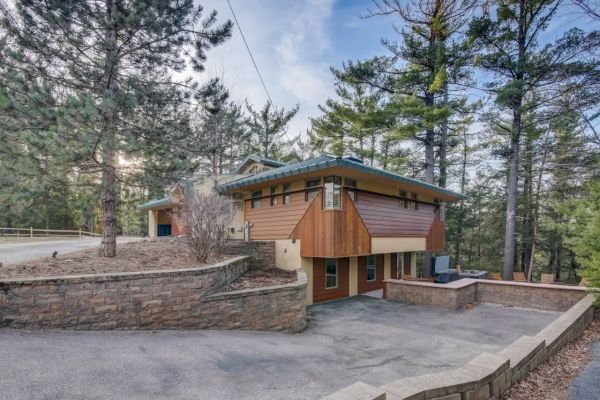 A modern A-frame style house with a metal roof, stone retaining walls, and a driveway surrounded by trees.