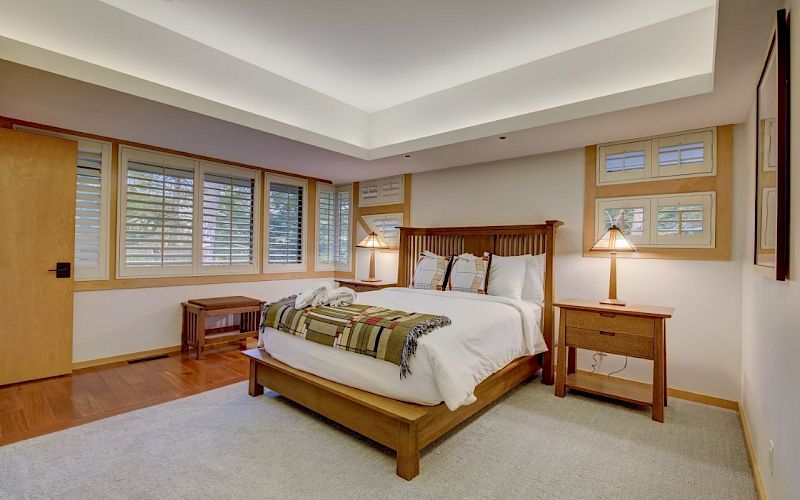 A bright bedroom with a wooden bed, nightstands, lamps, and large windows with blinds.