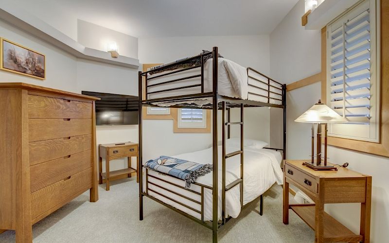 A tidy bedroom with bunk beds, a wooden dresser, a lamp, and a window with shutters.