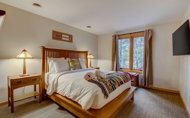 A tidy bedroom with a wooden bed, bedside lamp, and curtains framing a window with trees outside.