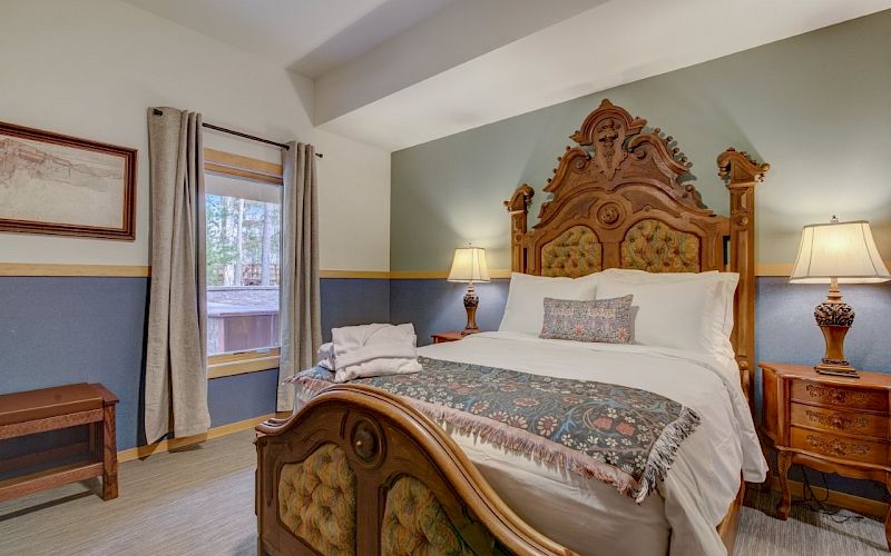 A furnished bedroom with a carved wooden bed, bedside lamps, framed art, and curtains near a window.