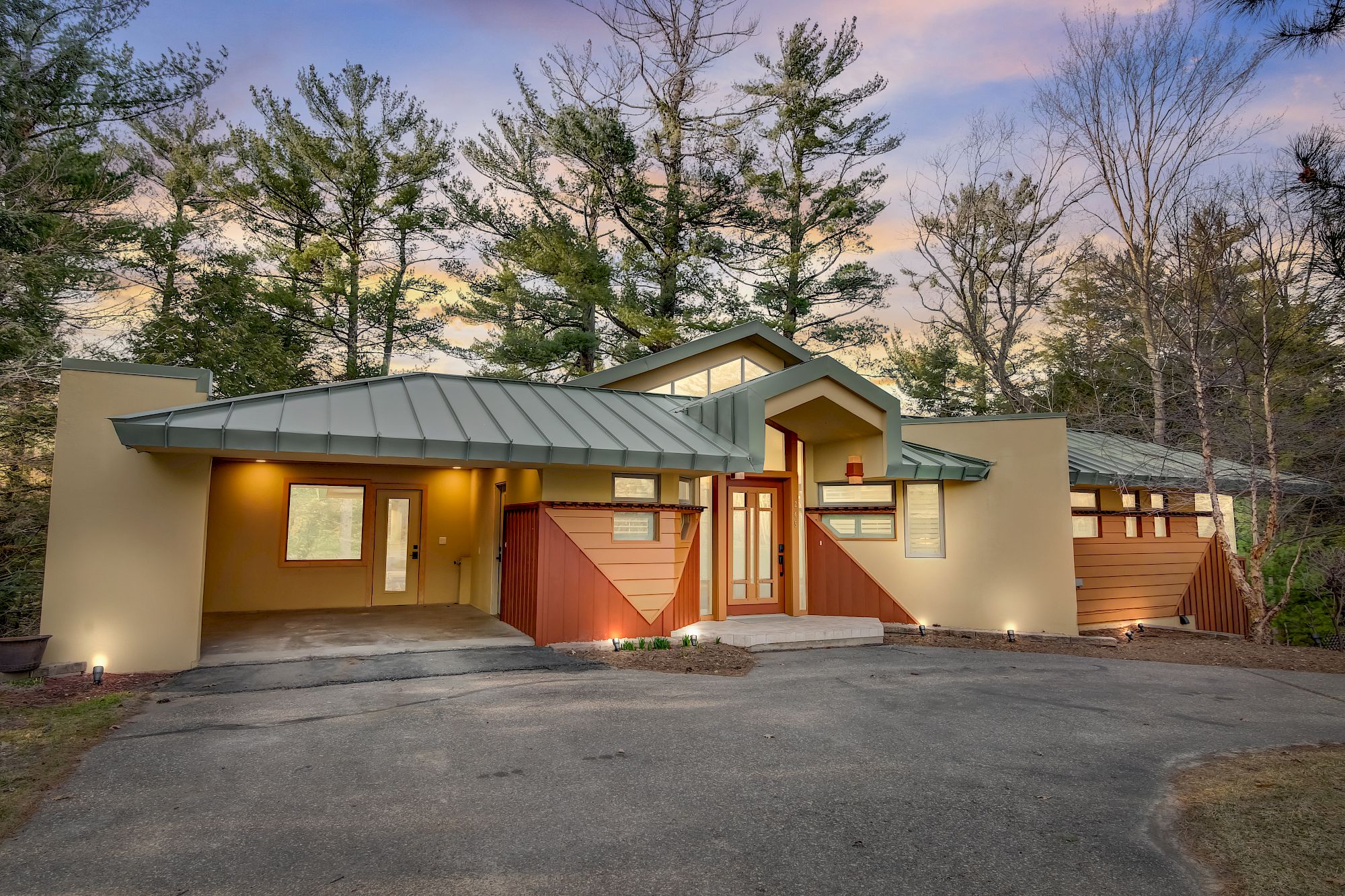 A modern house with a green metal roof, warm lights, and a red gate, set among trees at sunset.