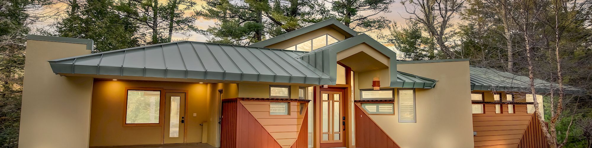 A modern house with a green metal roof, warm lights, and a red gate, set among trees at sunset.