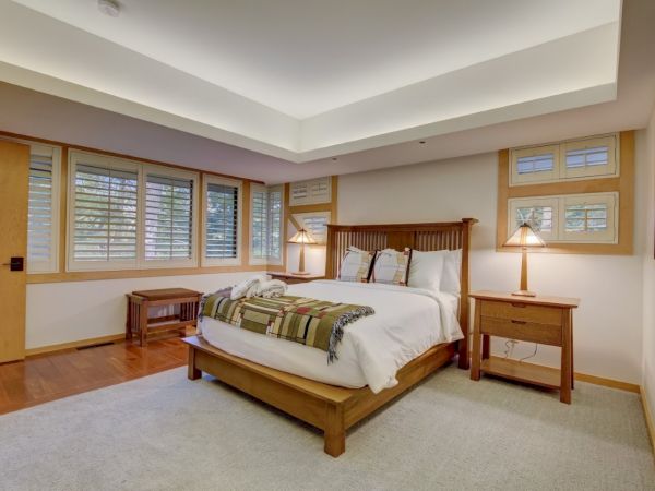 A cozy bedroom with a wooden bed, pillows, bedside tables, two lamps, and large windows with shutters.