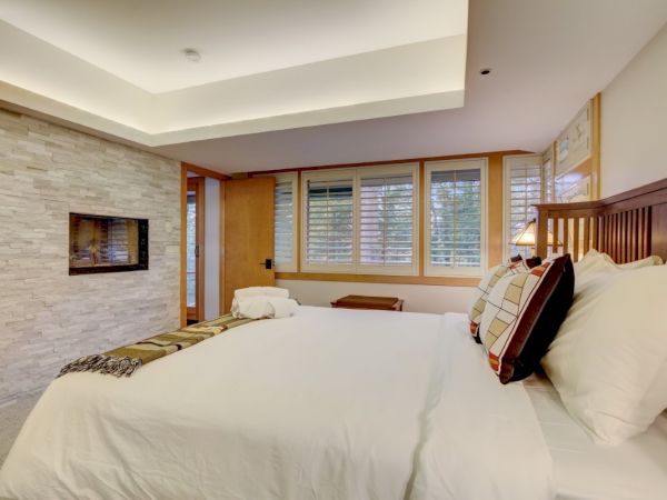 A modern bedroom with a large bed, stone fireplace wall, and wide windows with blinds, plus pillows and a wooden headboard.
