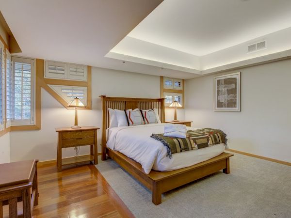 A bright bedroom with a wooden bed, decorative pillows, bedside lamps, windows, and framed art on clean white walls.