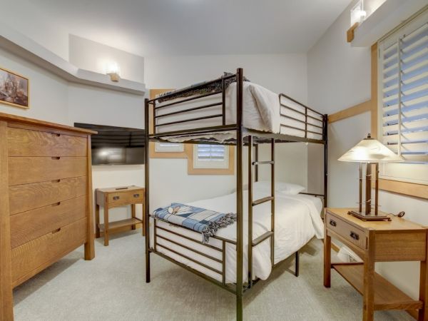 A cozy bedroom with a metal bunk bed, wooden dresser and nightstand, lamp, blinds, and framed wall art.