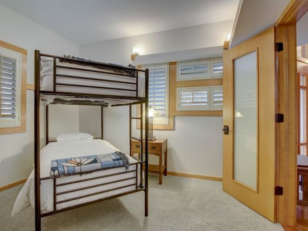 A bright bedroom with a metal bunk bed, bedside cabinet, and two windows with blinds and wall lights.