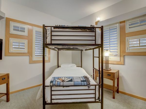 A small bedroom with a metal-framed bed, neatly made sheets and blanket, and windows with blinds plus a bedside table and lamp.