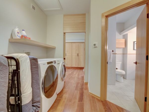 A bright laundry room with a washer/dryer, folded bedding, shelves with detergent, and a doorway to a tiled bathroom.