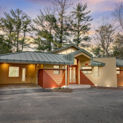 A modern home exterior with a green metal roof, warm-lit entrance, driveway, and pine trees under a pastel sunset.